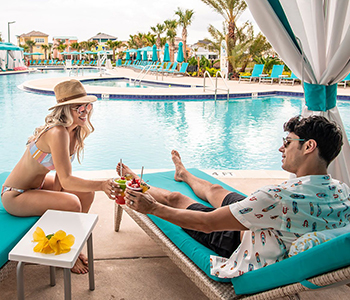 Casal tilintando copos em um brinde enquanto estão sentados em uma cabana privativa à beira da piscina do Margaritaville Resort Orlando.