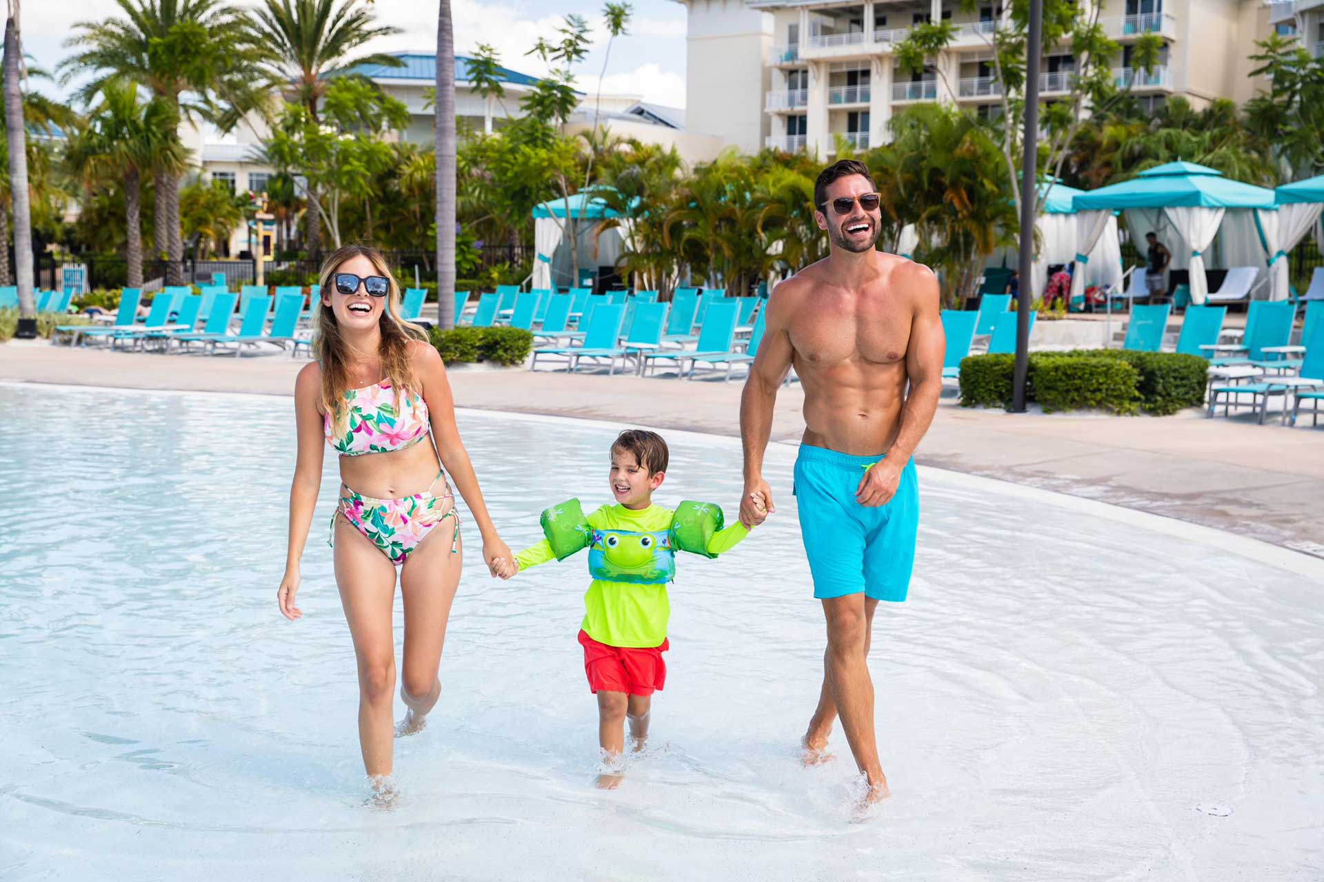 Родители гуляют с маленьким мальчиком в мелкой части бассейна курортного отеля Margaritaville Orlando.