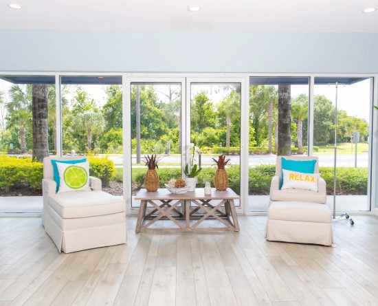 Entspannende Loungesessel und ein dekorativer Beistelltisch vor einem großen Fenster lassen natürliches Licht in die Vitamin Therapy Services Lounge im Margaritaville Resort Orlando.
