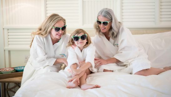 Grandmother, mother, and daughter wearing sunglasses and bath robes in their Margaritaville Resort Orlando room.